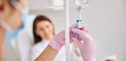 A closeup of PPE-gloved hands adjusting an infusion bottle with a smiling patient in the background.